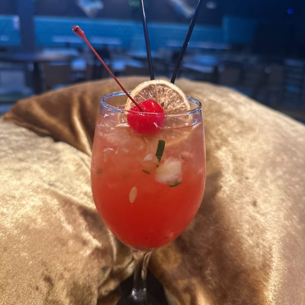 Vibrant red cocktail with a cherry, dried orange, and cucumber, resting on a golden cushion.