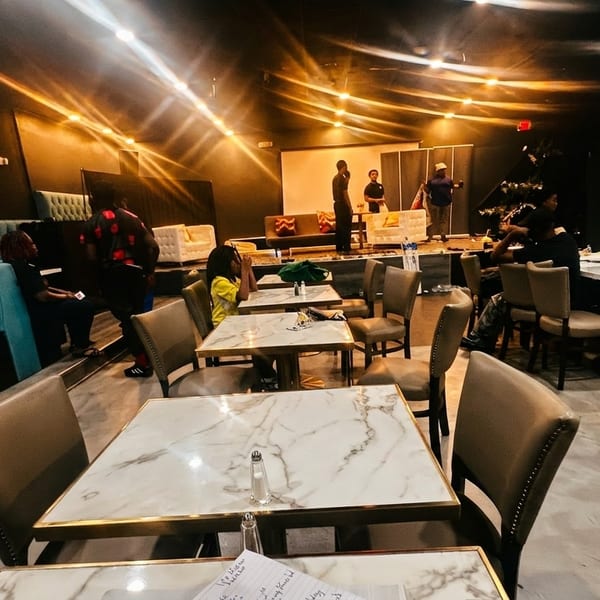 A dark restaurant interior with marble tables, teal booths, and people gathered around a stage.