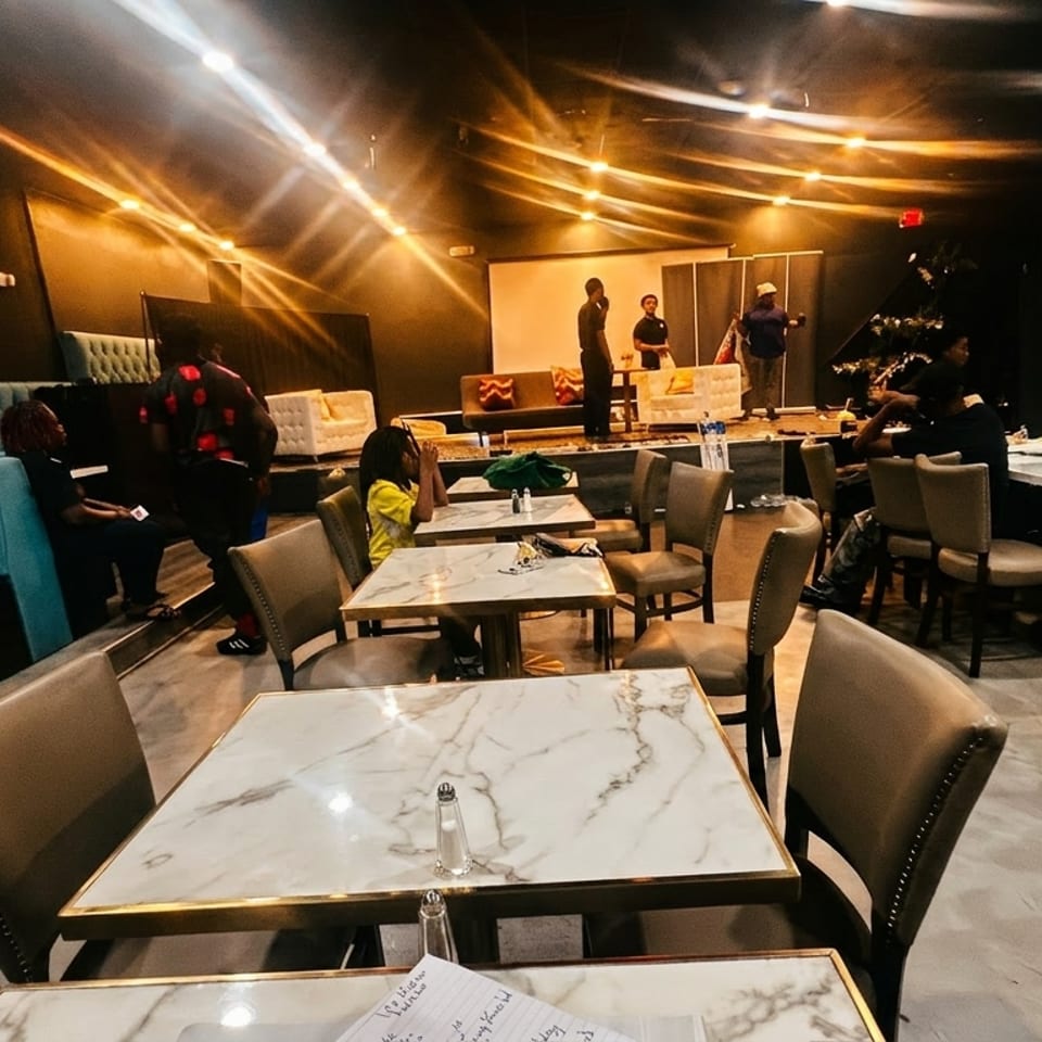 A dark restaurant interior with marble tables, teal booths, and people gathered around a stage.